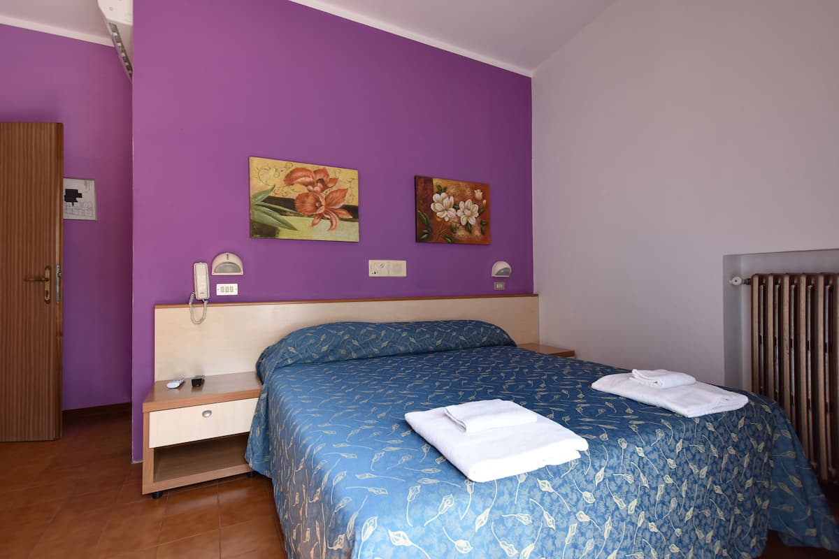 A bedroom with a purple wall, a bed with a blue floral bedspread, a bedside table with a lamp, and a wall-mounted phone.