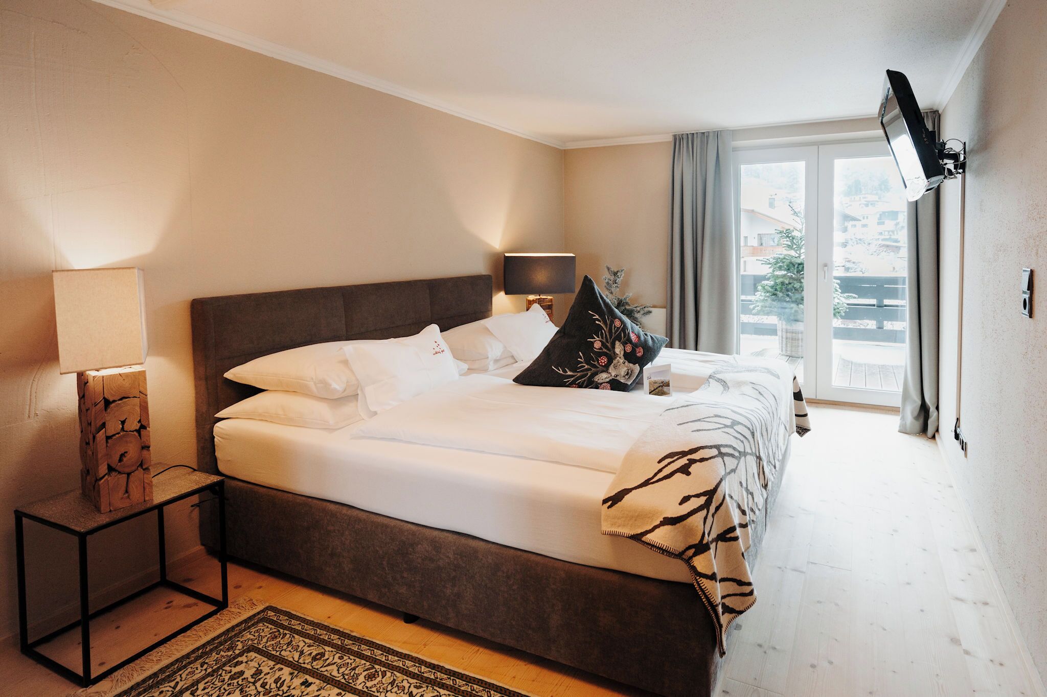 Un cuarto de hotel con una cama grande, una mesita de noche con lámpara, un televisor montado en la pared y vista al exterior a través de una ventana.