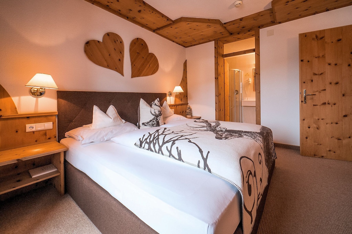 Ein Hotelzimmer mit einem großen Bett, einem Holzkopfstück, zwei Nachttischen mit Lampen, einer Holztür und einem Duschbereich.