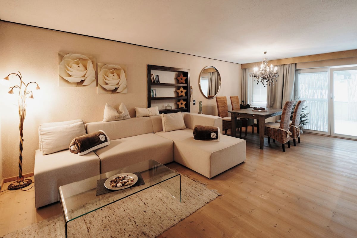 Ein modernes Wohnzimmer mit einem Ecksofa, einem Glastisch, einem Essbereich mit einer Kronleuchter und einem großen Fenster.