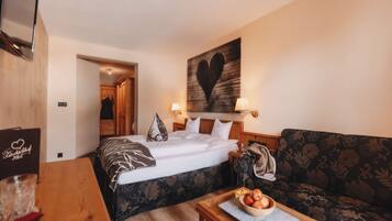A hotel room with a bed, a sofa, a wooden table with a bowl of fruit, and a wall with a heart-shaped artwork.