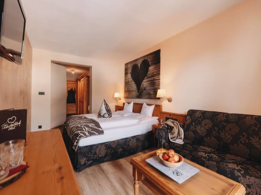 A hotel room with a bed, a sofa, a wooden table with a bowl of fruit, and a wall with a heart-shaped artwork.