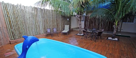 Una piscina exterior con una cubierta de madera, rodeada de sillas y una mesa, y una valla de bambú al fondo.