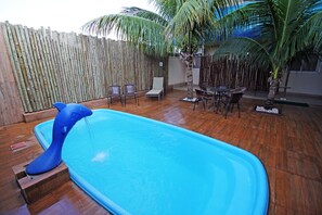 An outdoor swimming pool with a wooden deck, surrounded by chairs and a table, and a bamboo fence in the background.