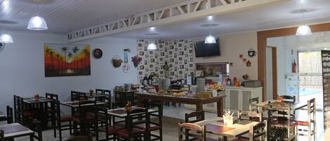 Un restaurante con varias mesas y sillas, una zona de cocina y un televisor en la pared.