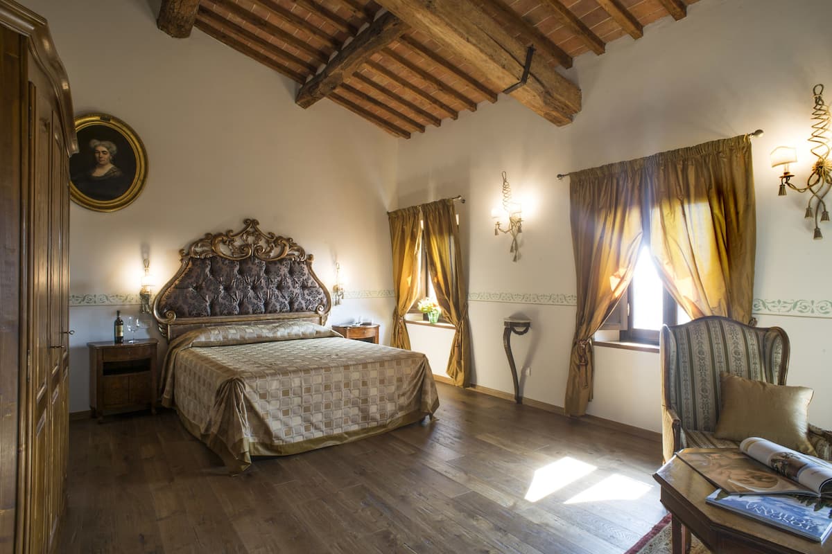 Un dormitorio con una cama grande, una silla, una mesita de noche, una lámpara, un cuadro en la pared y una ventana con cortinas.