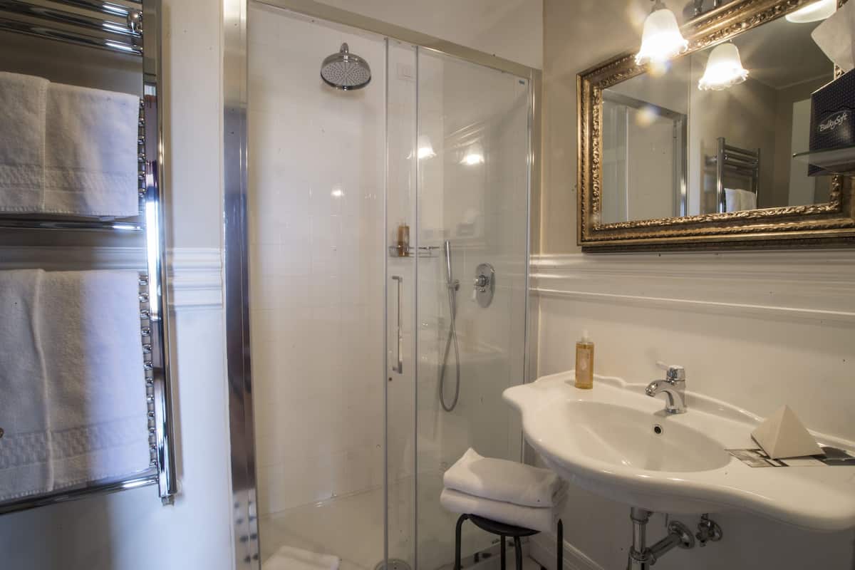Un baño con ducha acristalada, un lavabo blanco con un espejo encima y un perchero con toallas blancas.