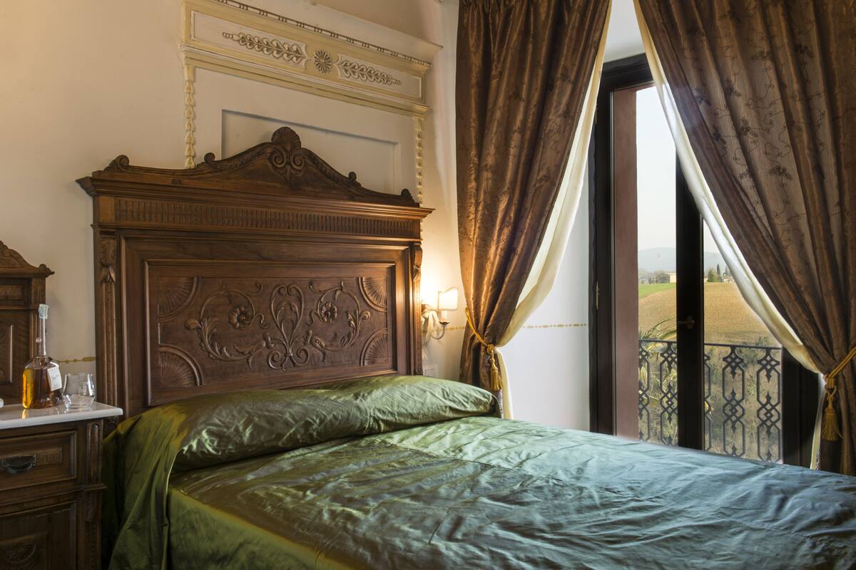 Una habitación con cabecera de madera, cama con ropa de cama verde, mesita de noche con una botella y vasos, y vista a través de una ventana con cortinas.