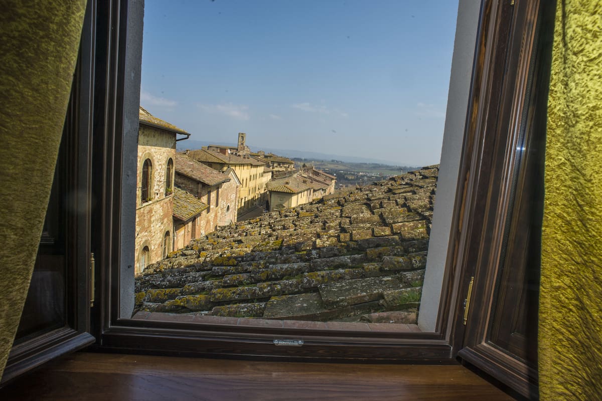 Vista a través de una ventana de una escalera de piedra que conduce a un edificio con ventanas de arco y una torre, sobre un paisaje montañoso.