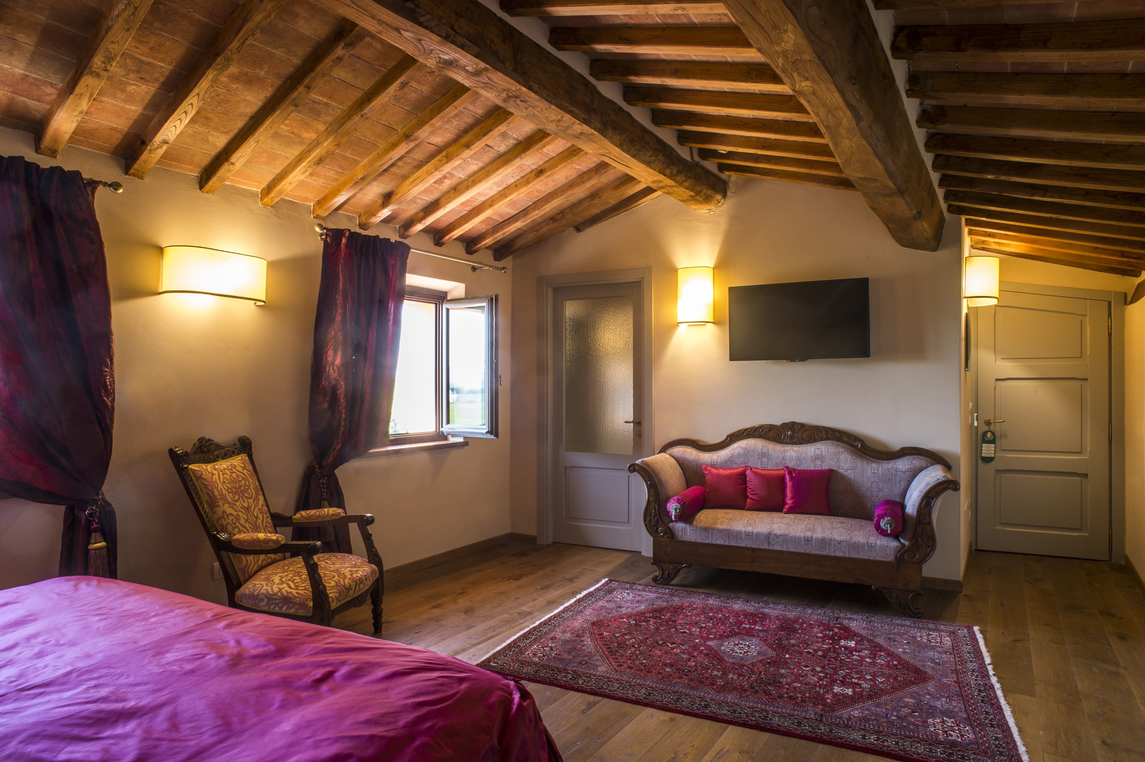 A room with a wooden ceiling, a sofa with cushions, a chair, a bed, and a wall-mounted TV.