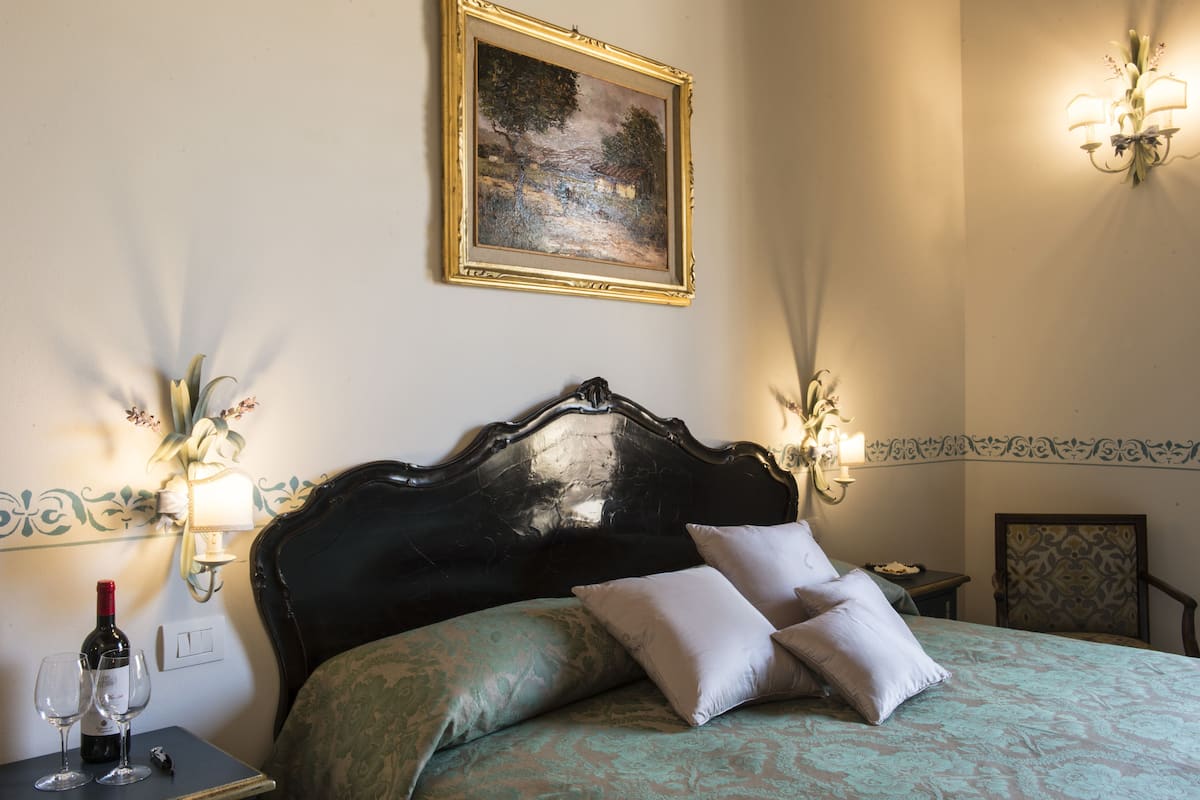 Un dormitorio con cama, una mesita de noche con copas de vino, un cuadro en la pared y un aplique.