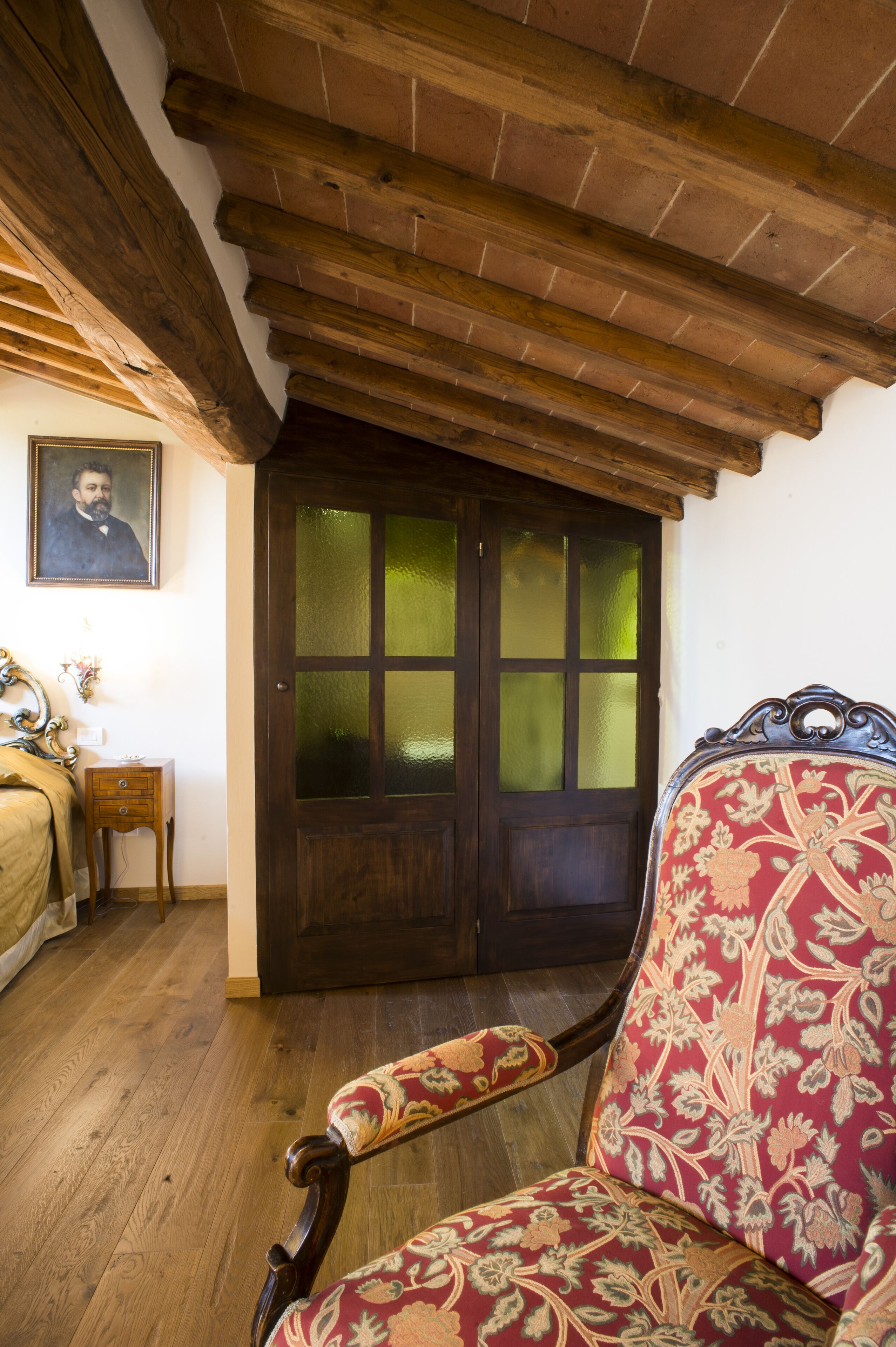 A room with a wooden ceiling, a dark wooden door with glass panels, a floral-patterned armchair, and a small wooden nightstand with a lamp.