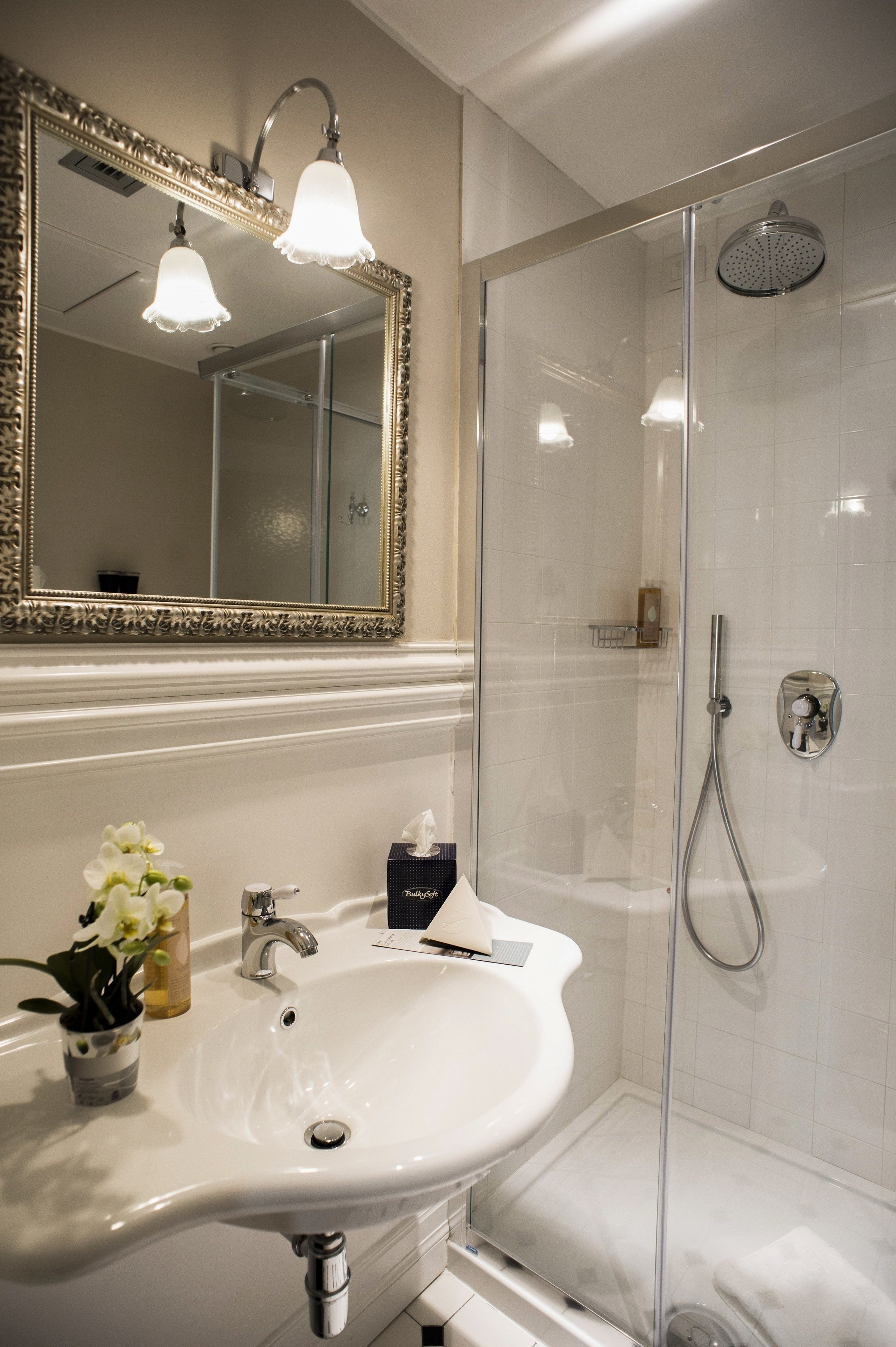 A bathroom with a white sink, a mirror with ornate framing, a shower with a glass door, and a towel rack.