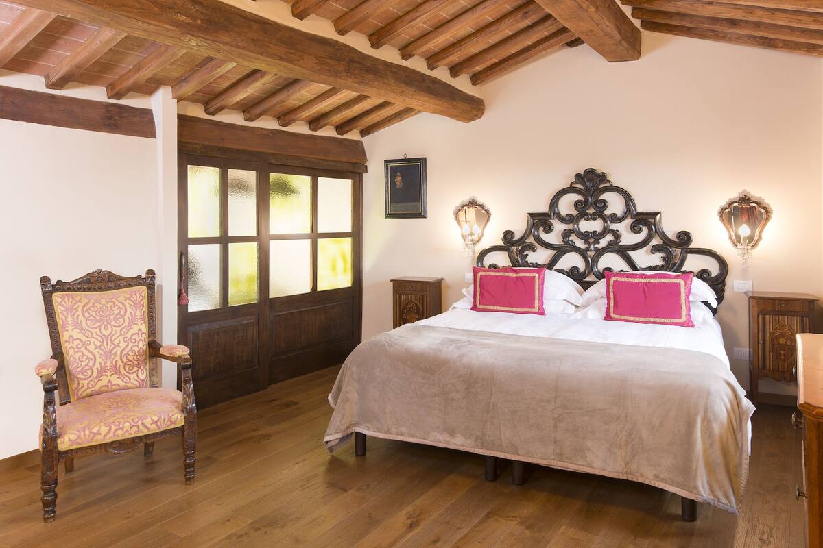 Un dormitorio con techo de madera, una cama con cabecera, dos mesitas de noche, una silla y un ventanal con cristal esmerilado.