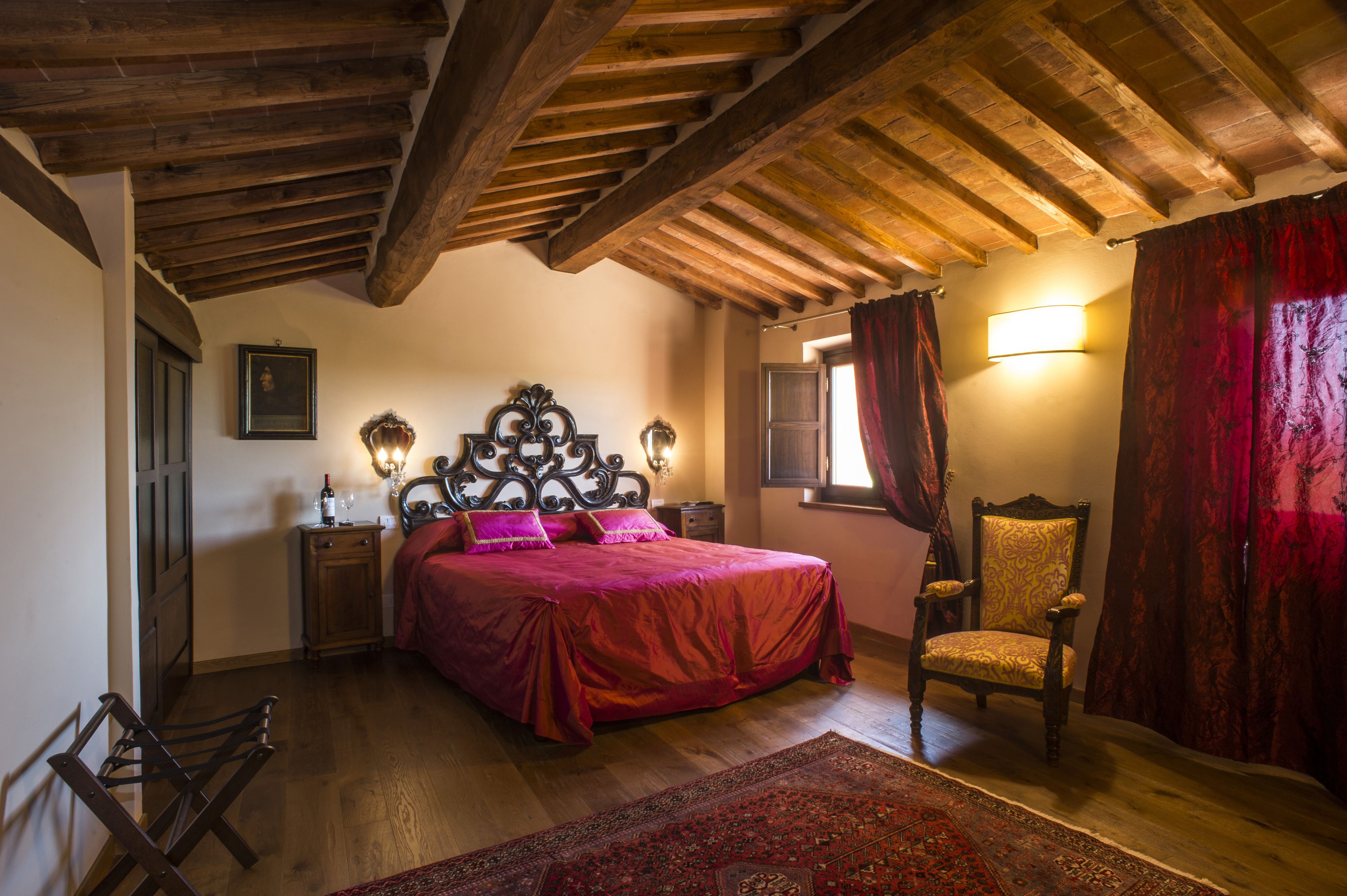 Una camera da letto con soffitto in legno, un letto con copripiumino rosso, una sedia, un comodino con una lampada e una finestra con tende.