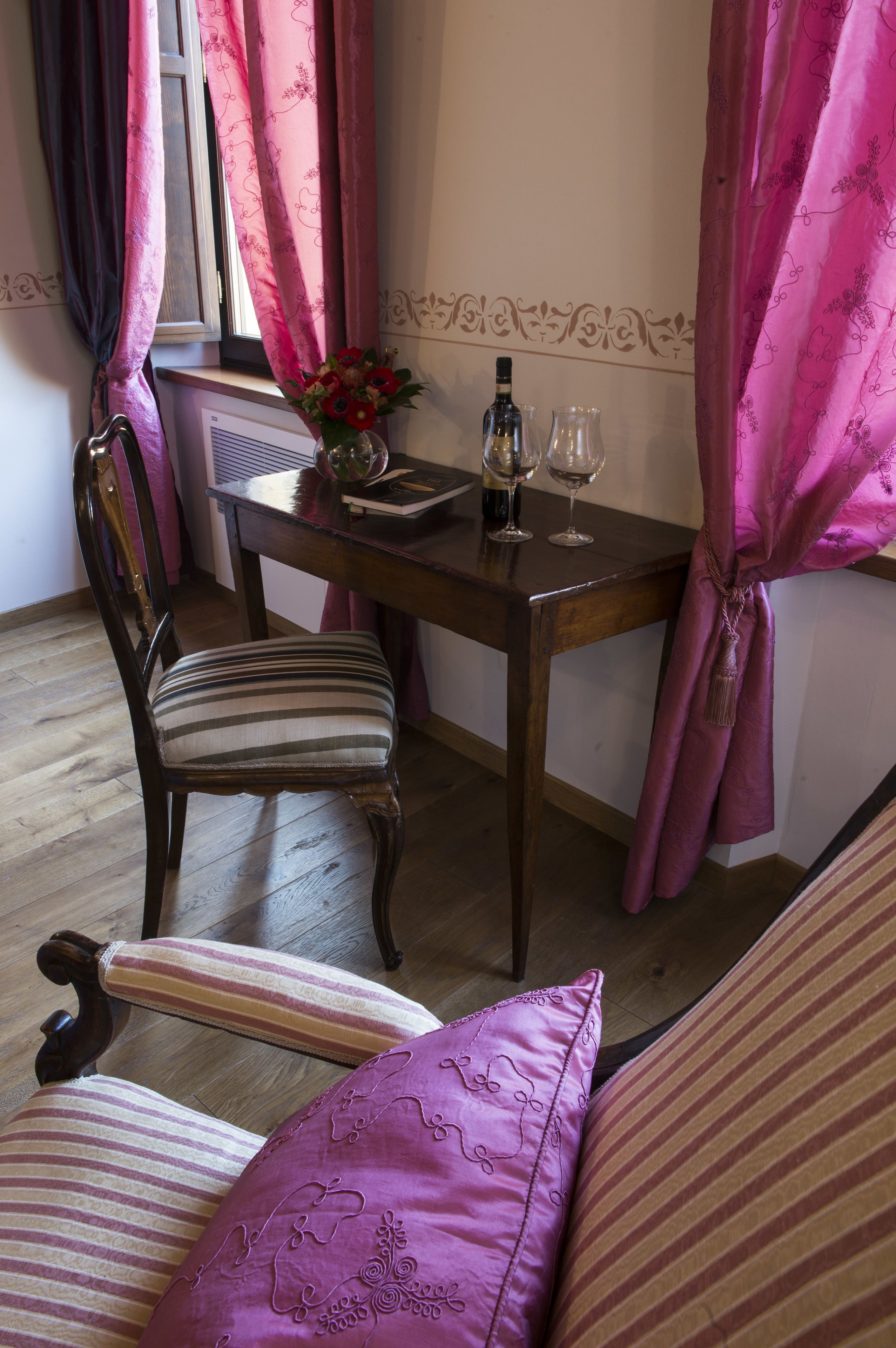A dining area with a wooden table, a chair, and a sofa. There is a window with pink curtains, a vase with flowers, and two wine glasses on the table.