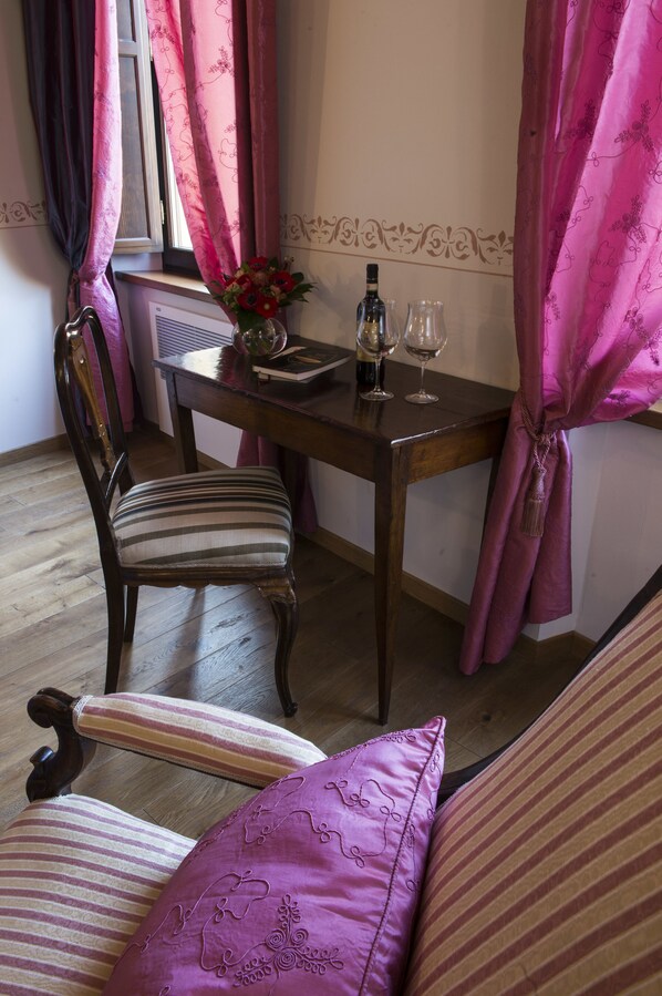 A dining area with a wooden table, a chair, and a sofa. There is a window with pink curtains, a vase with flowers, and two wine glasses on the table. - Palazzo Pacini (Colle di Val d'Elsa)