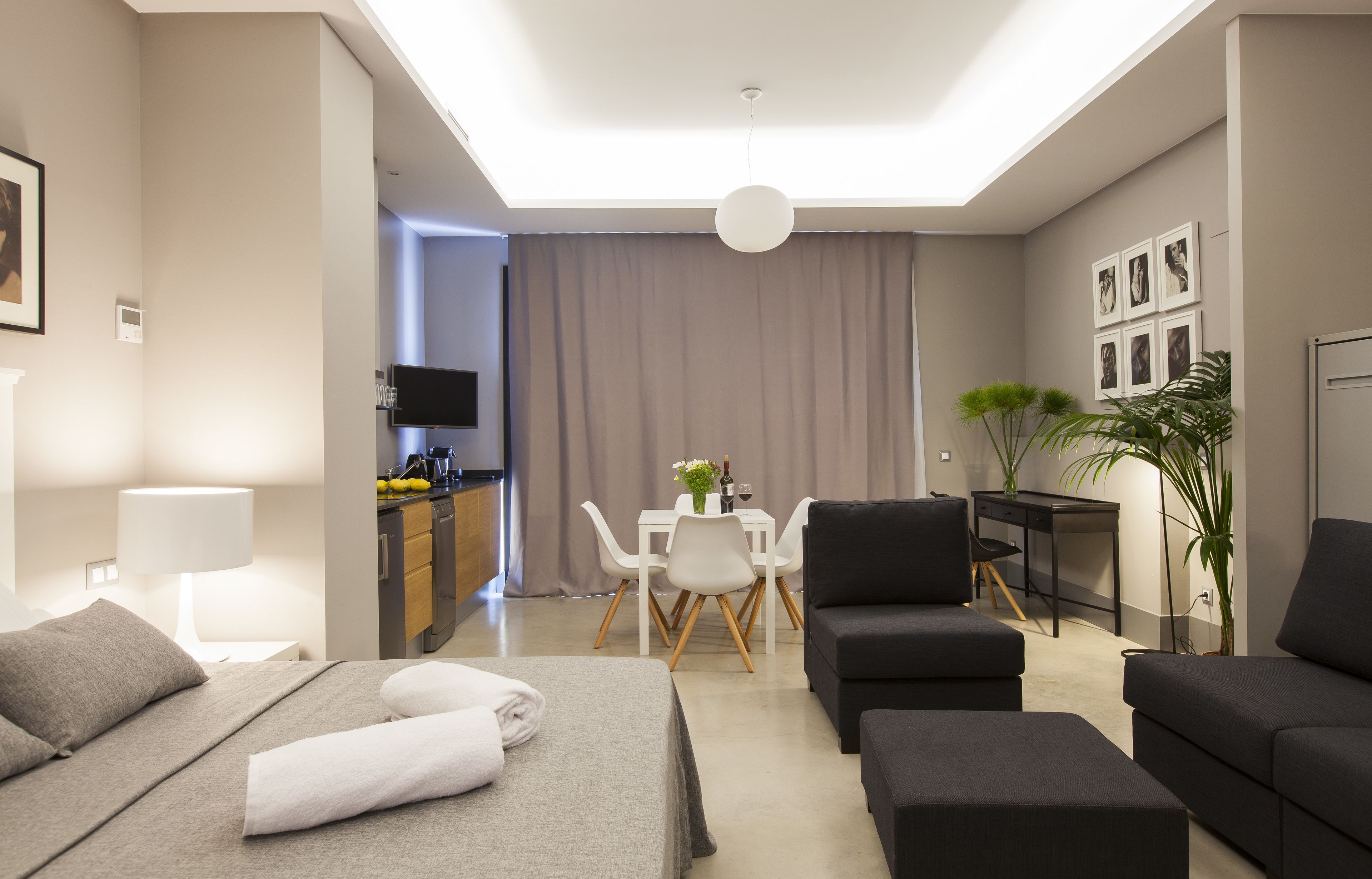 A modern hotel room with a bed, a sofa, a dining table with chairs, a desk, and a TV.