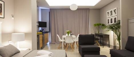 A modern hotel room with a bed, a sofa, a dining table with chairs, a desk, and a TV.