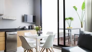 Une cuisine moderne équipée d’un lave-vaisselle, d’une table à manger avec des chaises blanches et d’une grande fenêtre donnant sur un balcon.