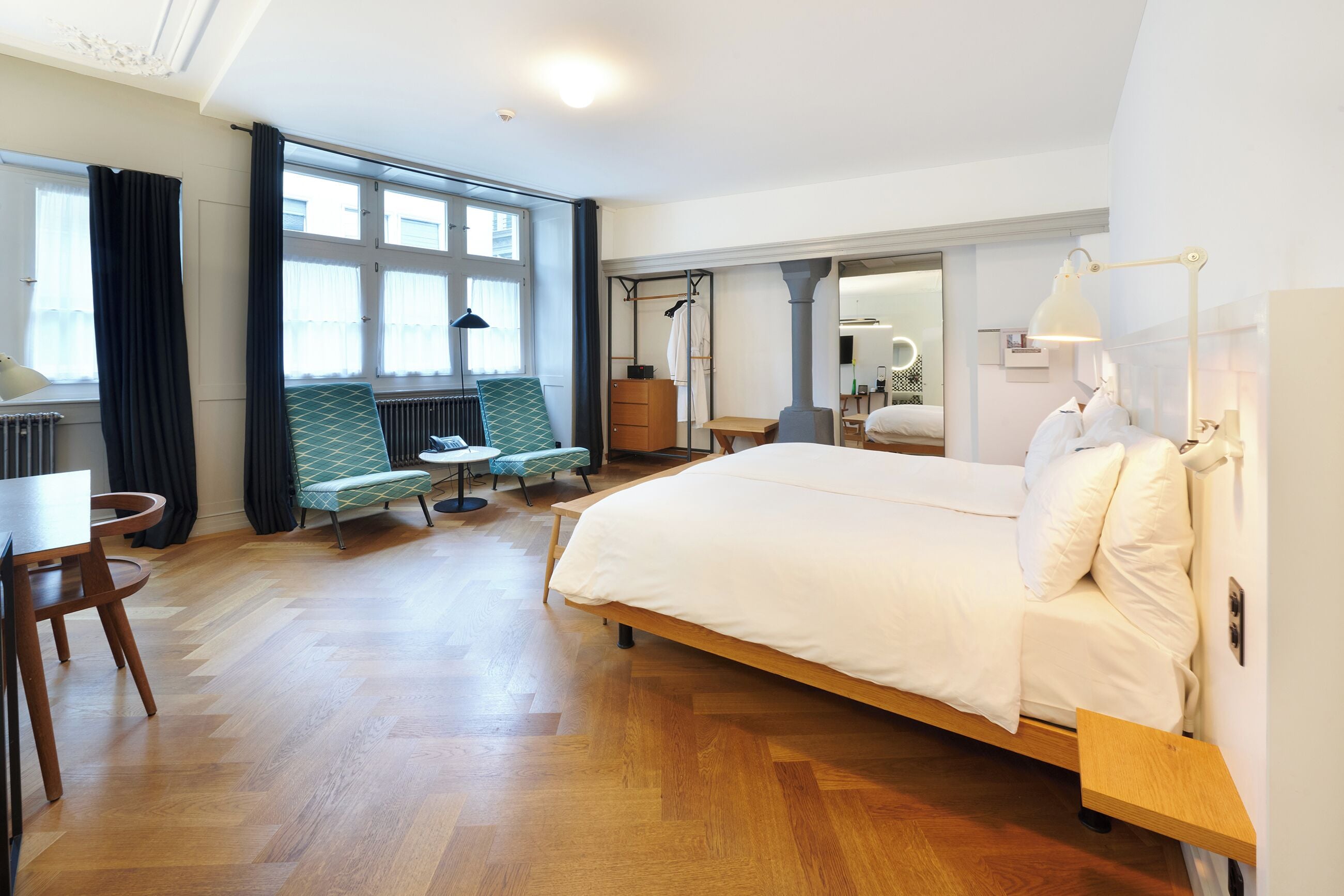 Habitación de hotel con una cama grande, zona de estar con sofá y sillones, una mesita y una ventana con cortinas.