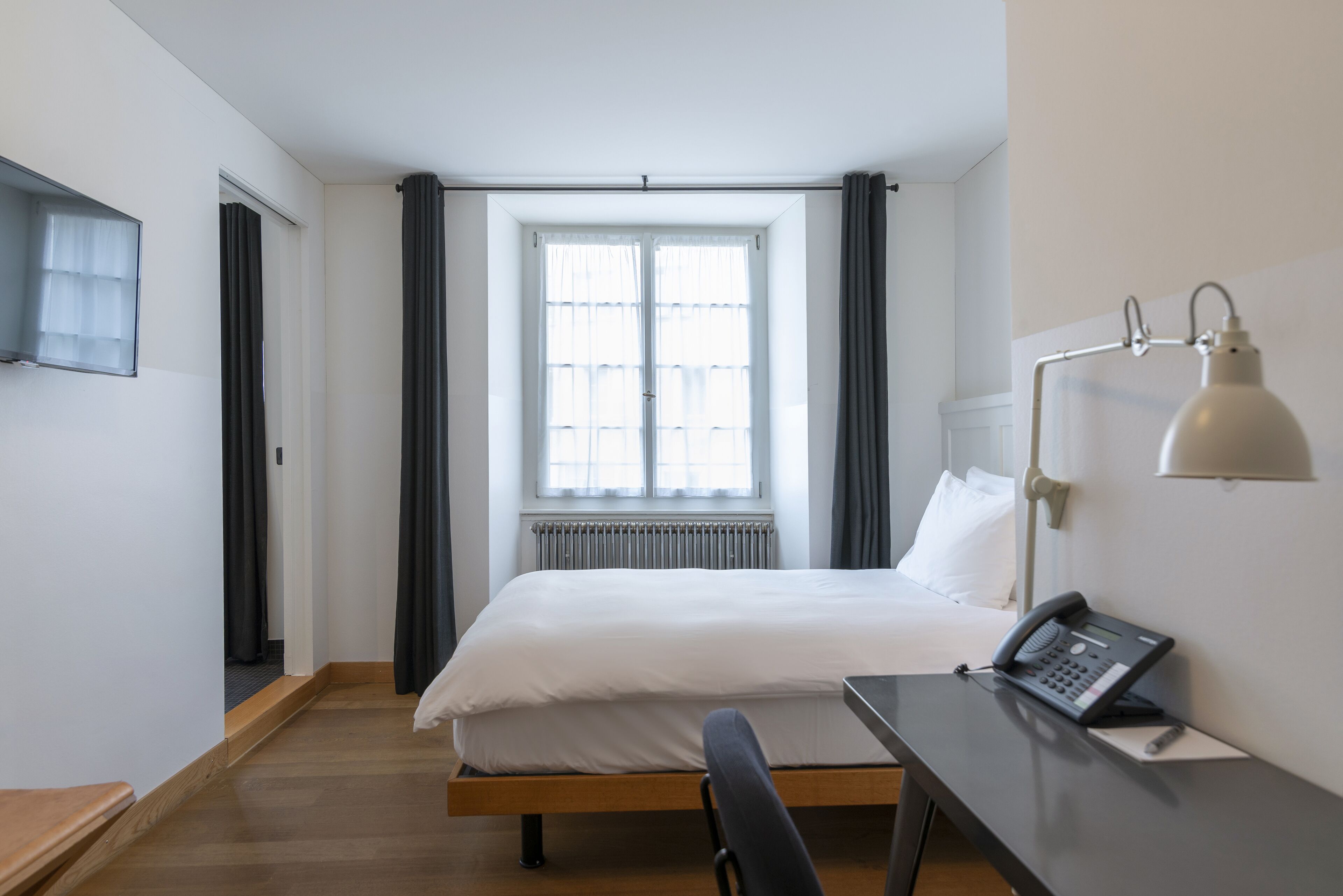A hotel room with a bed, a desk with a telephone, a lamp, and a window with curtains.