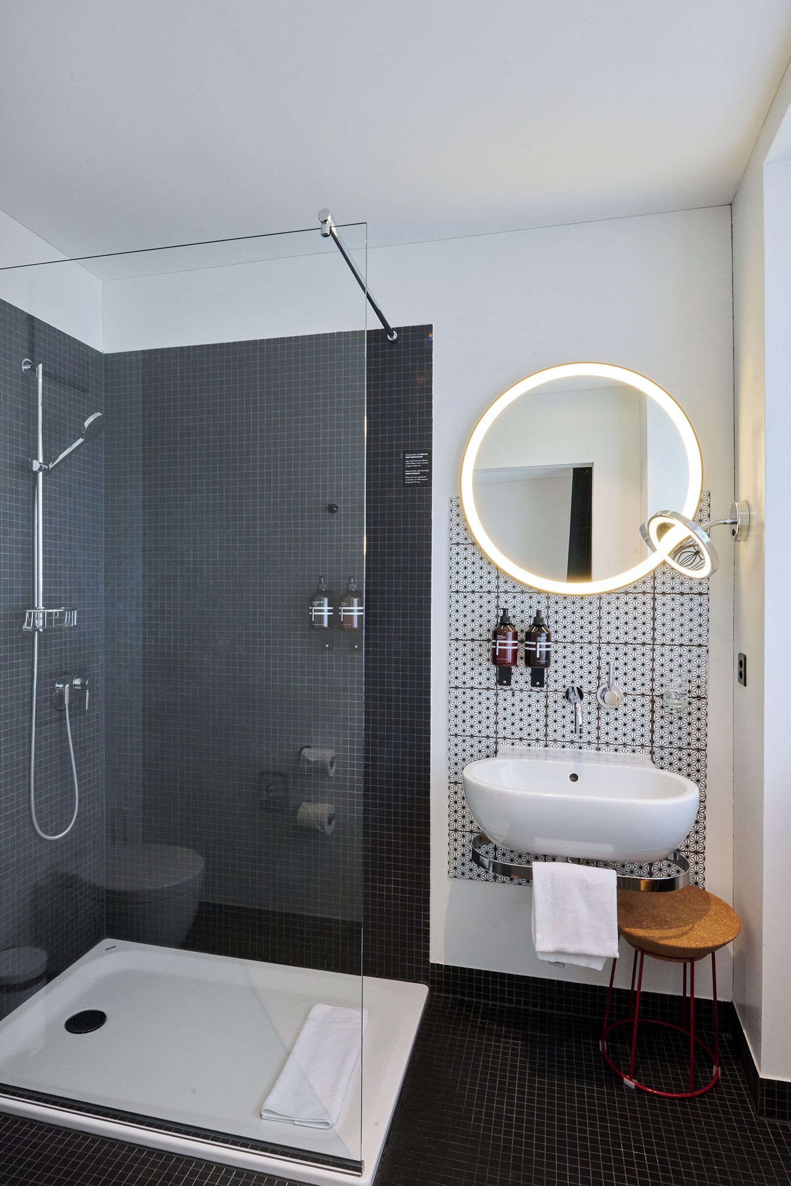 A modern bathroom with a glass-enclosed shower, a white sink with a mirror, and a small stool.