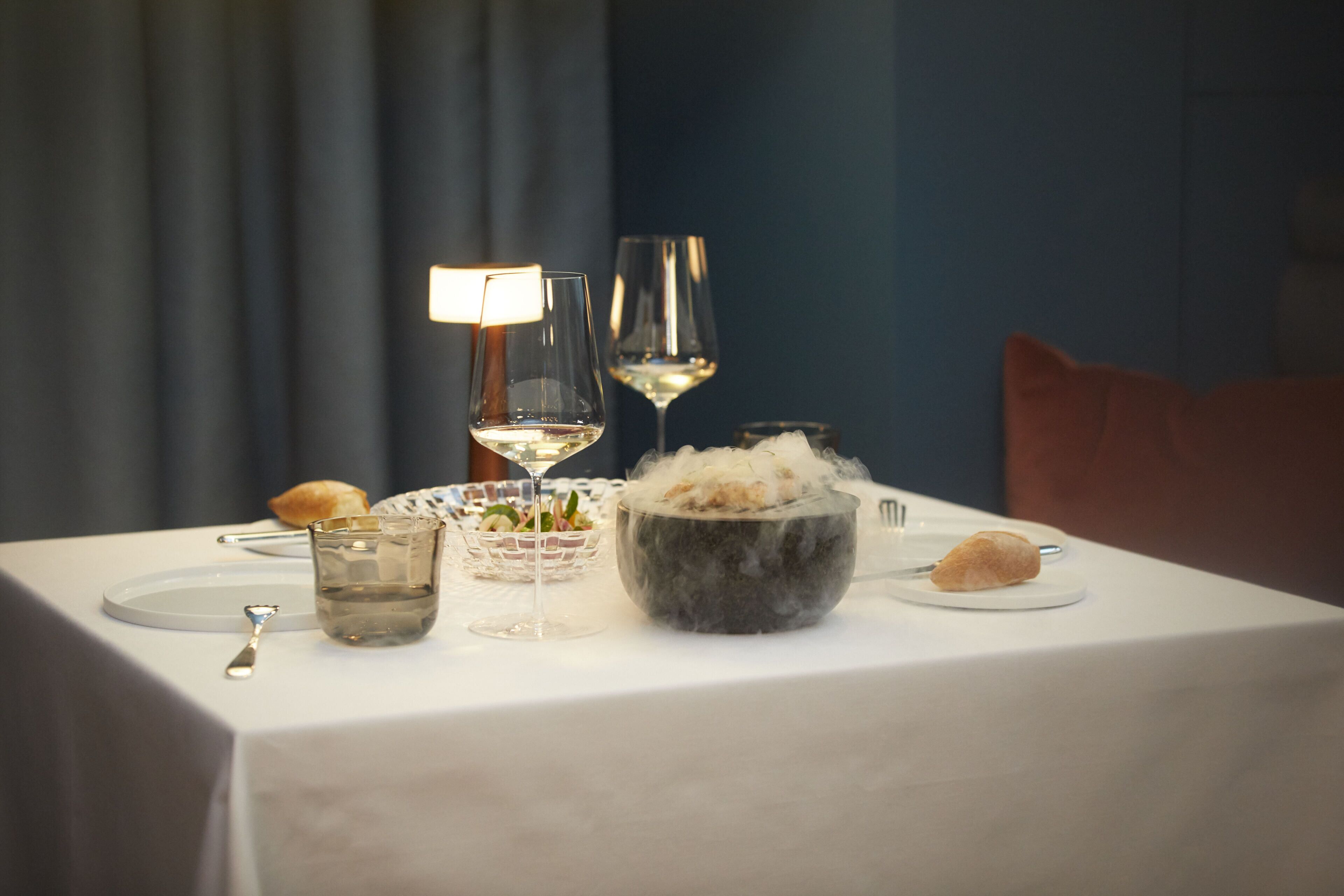Una mesa de comedor dispuesta con una vela, copas de vino, un tazón de helado y un croissant.