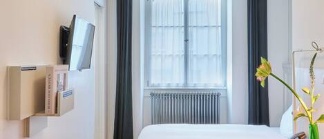 A hotel room with a bed, a desk, a chair, a TV mounted on the wall, a window with curtains, and a radiator.