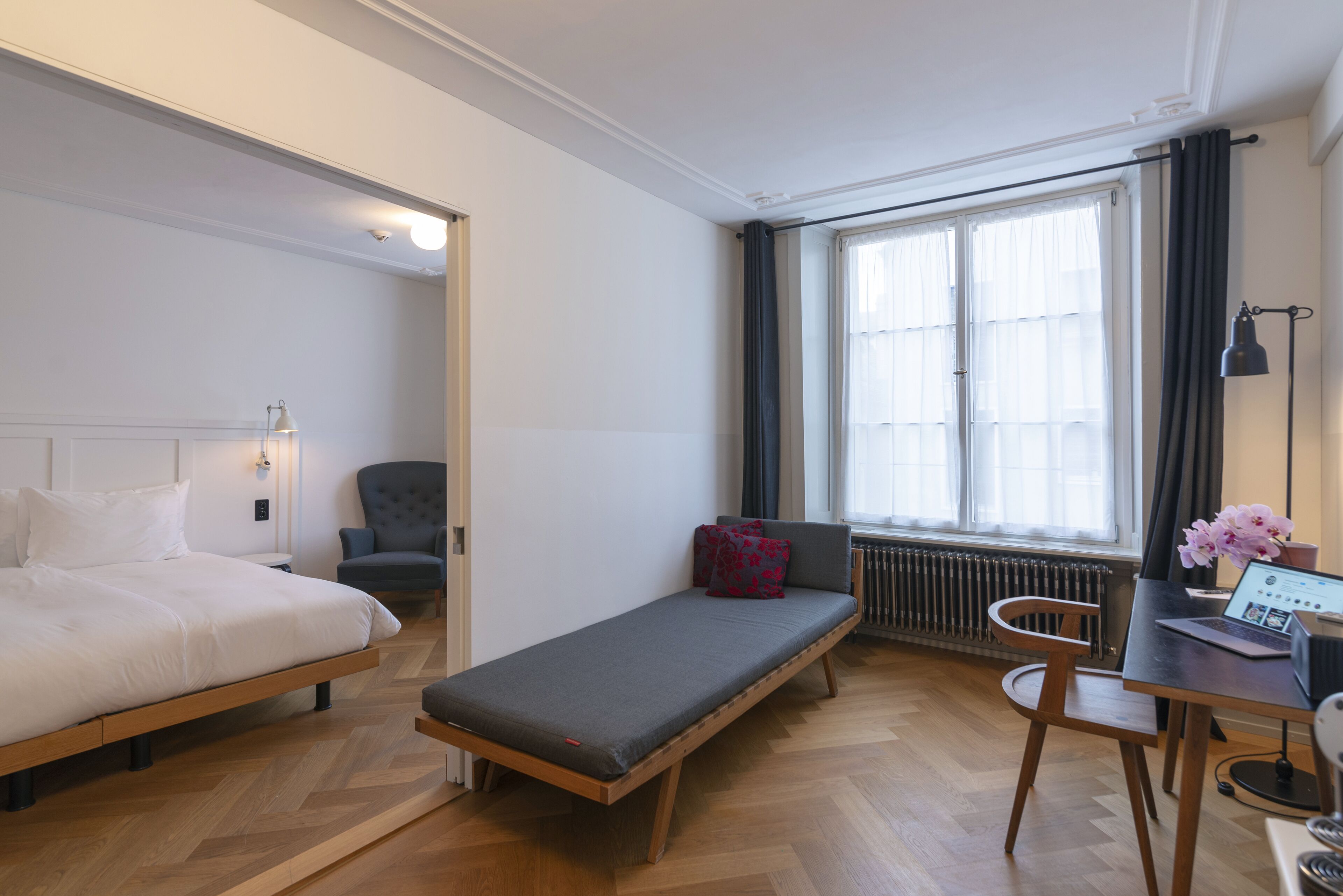 Una camera d'albergo moderna con un letto, un divanetto, una scrivania con un portatile e una finestra con tende.