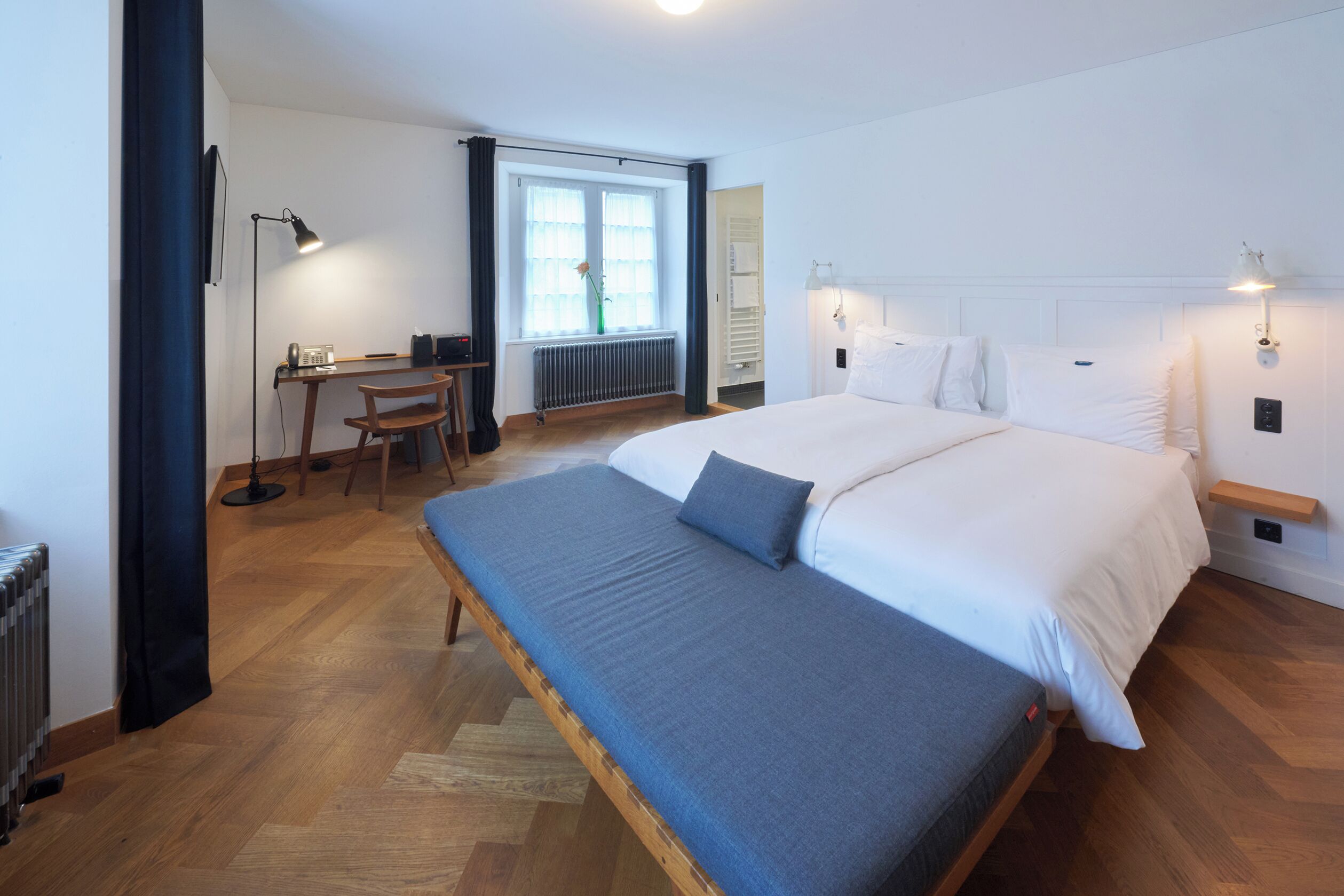 Ein Schlafzimmer mit einem großen Bett, einem Schreibtisch und einem Stuhl. Es gibt ein Fenster mit Vorhängen, eine Heizung und eine Wandlampe.