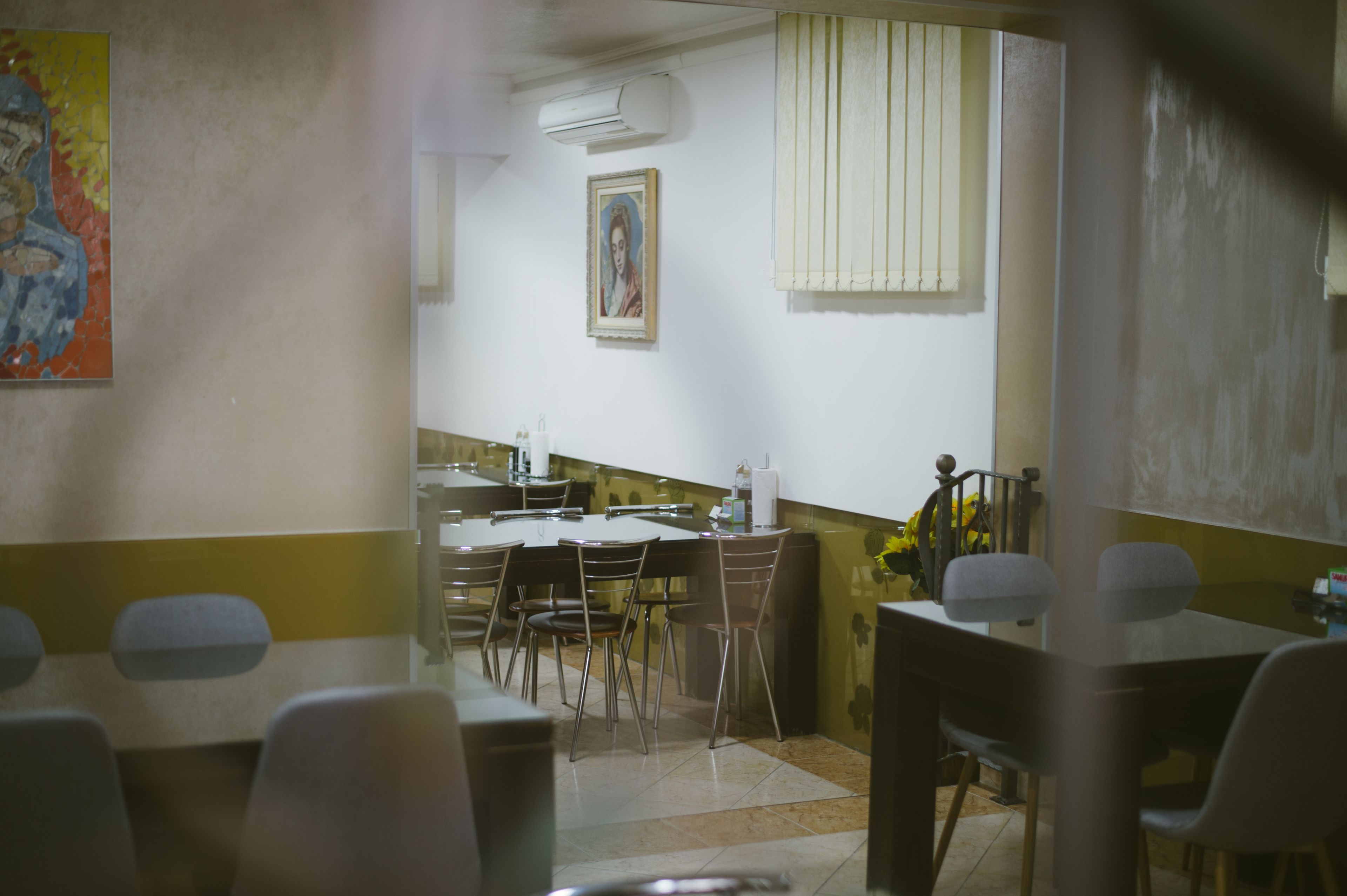 A dining area with tables and chairs, a painting on the wall, and a window with blinds.