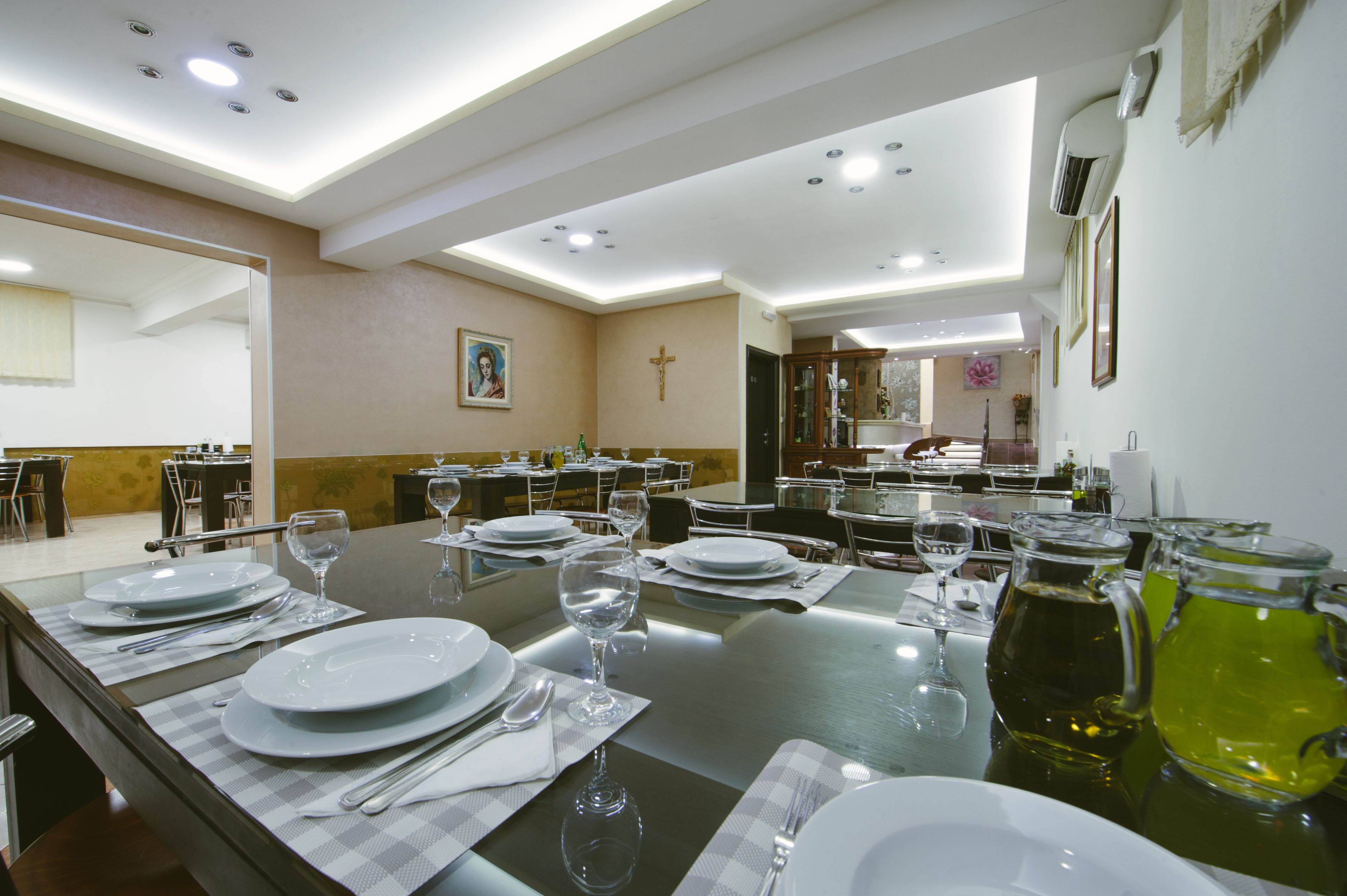 Une salle à manger avec des tables dressées pour le service, une croix accrochée au mur et une vue sur la pièce adjacente.