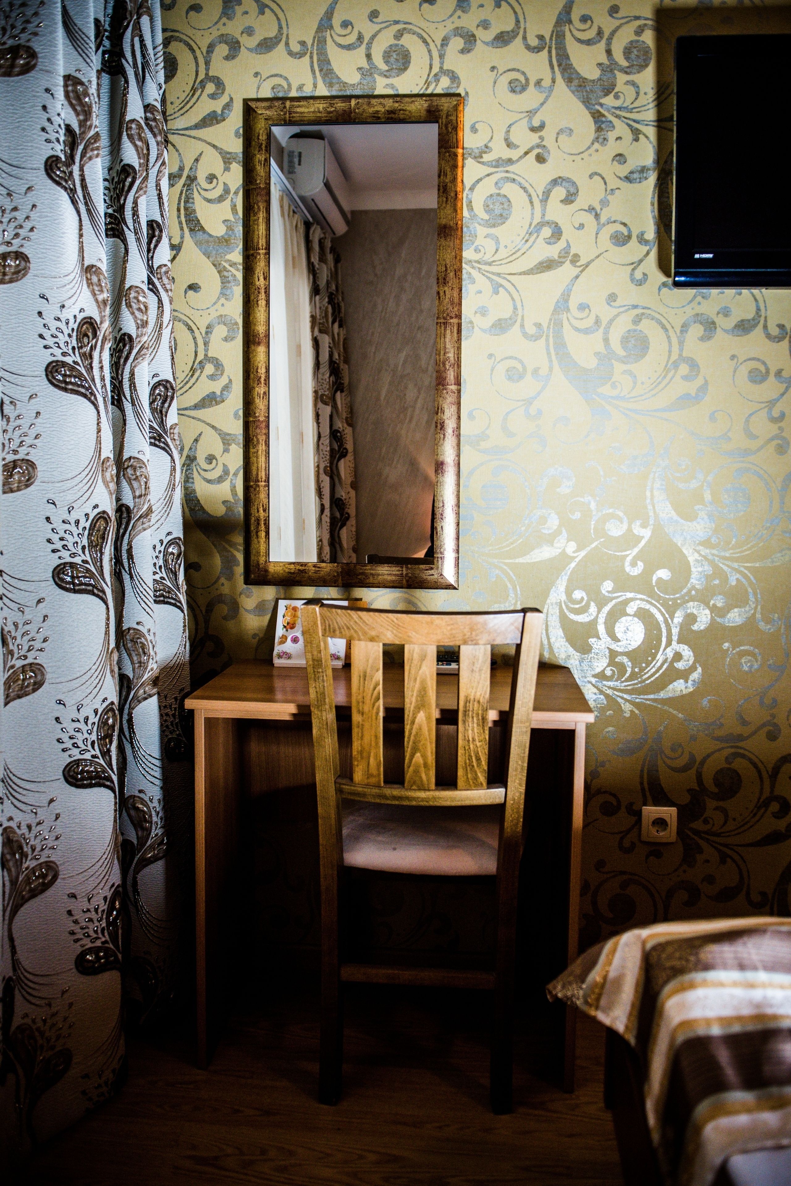 A room with a wooden desk, a chair, a mirror, a patterned wall, and a curtain.