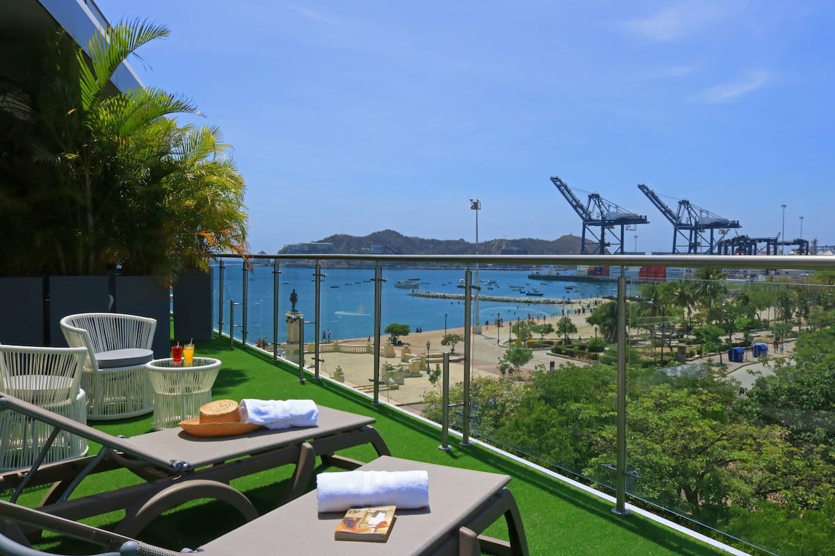 A balcony with outdoor seating, a view of the sea, and distant cranes.