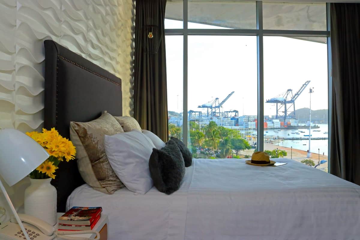 A hotel room with a large bed, a nightstand with a lamp and books, a vase with flowers, and a view of a port with cranes.