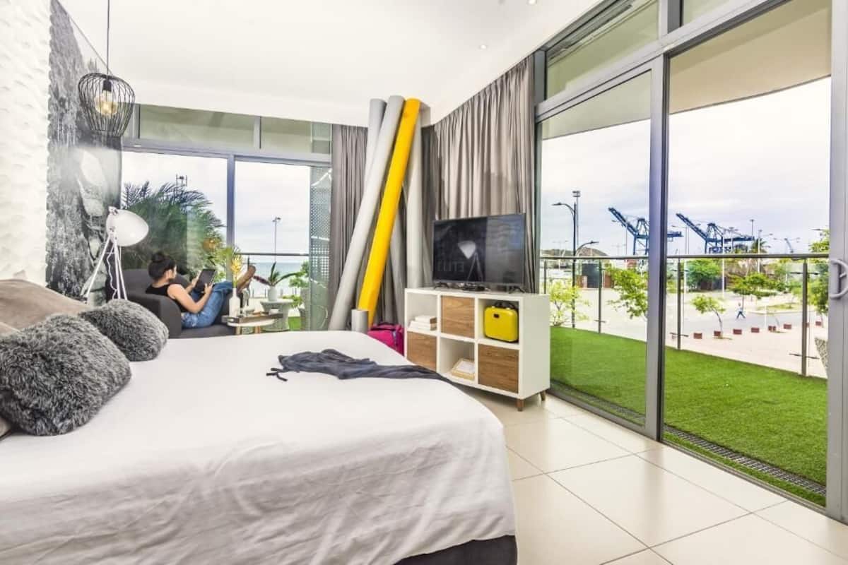 A modern hotel room with a large bed, a TV, and a balcony with a view of the port.