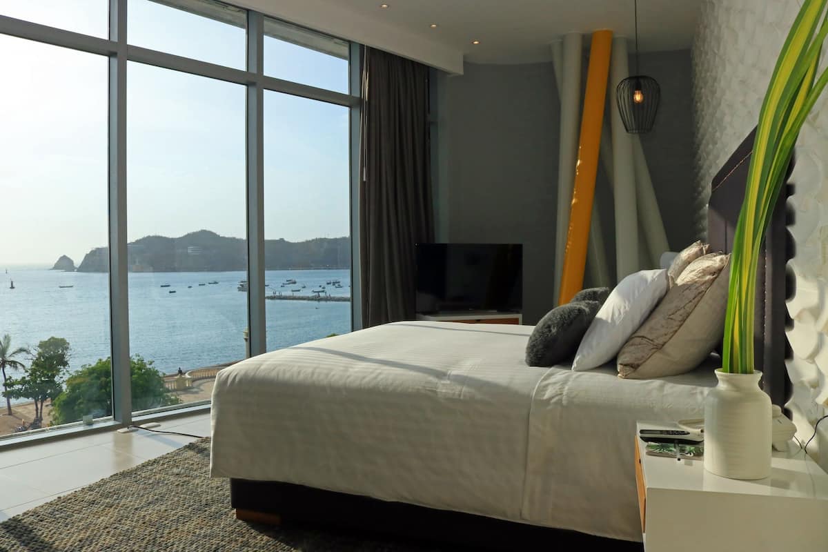 A bedroom with a large window overlooking a body of water, a bed, a television, and a bedside table with a vase.