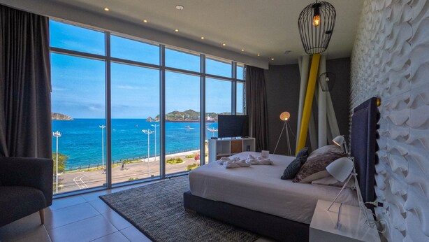 A bedroom with a large window overlooking a coastal view, a bed, a bedside table, and a modern lamp. - Bonita Bay Concept Hotel by Xarm Hotels (Santa Marta)
