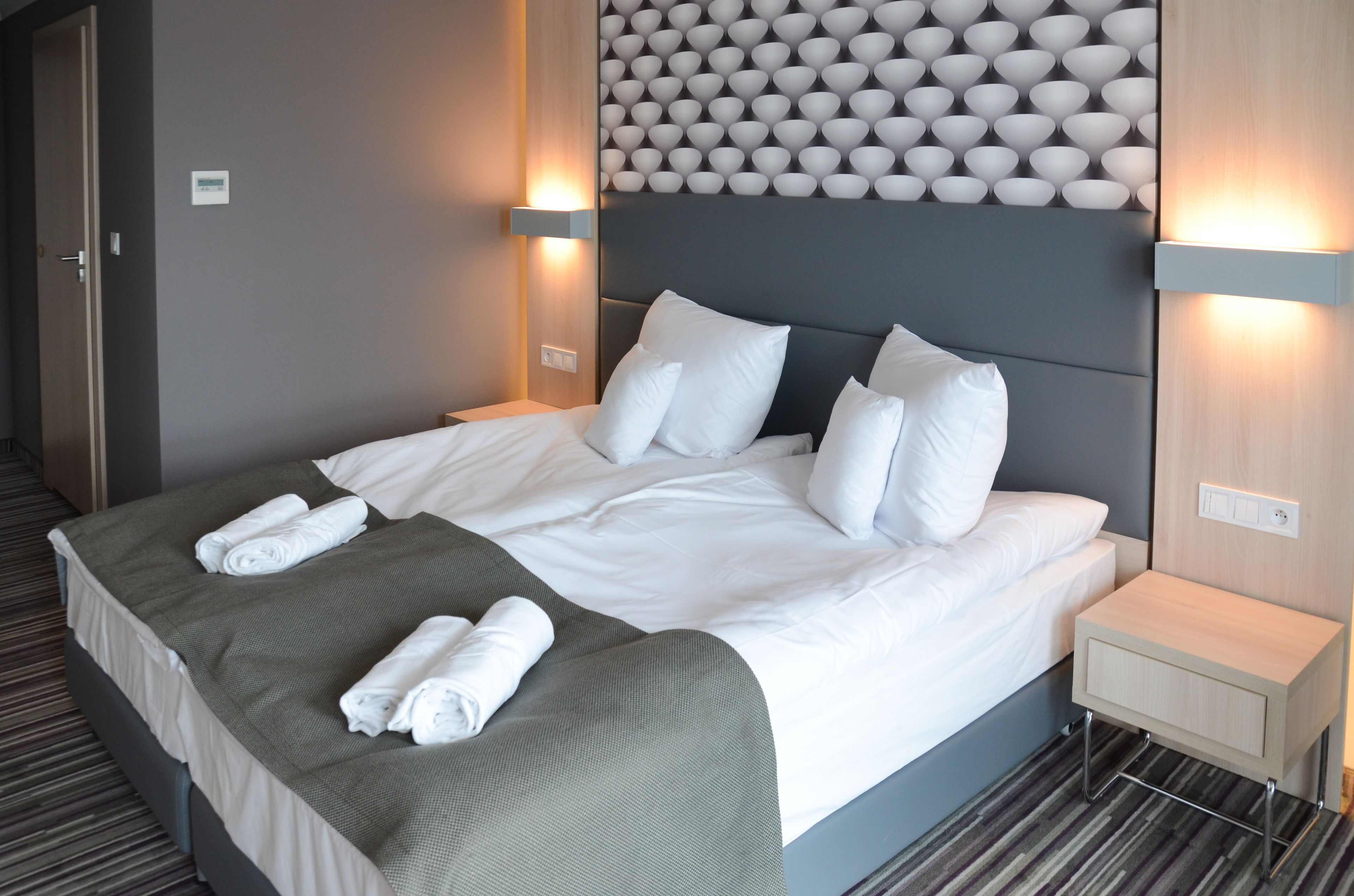 A neatly made bed with white linens and two rolled towels, a grey headboard, and two bedside tables with wall-mounted lamps.