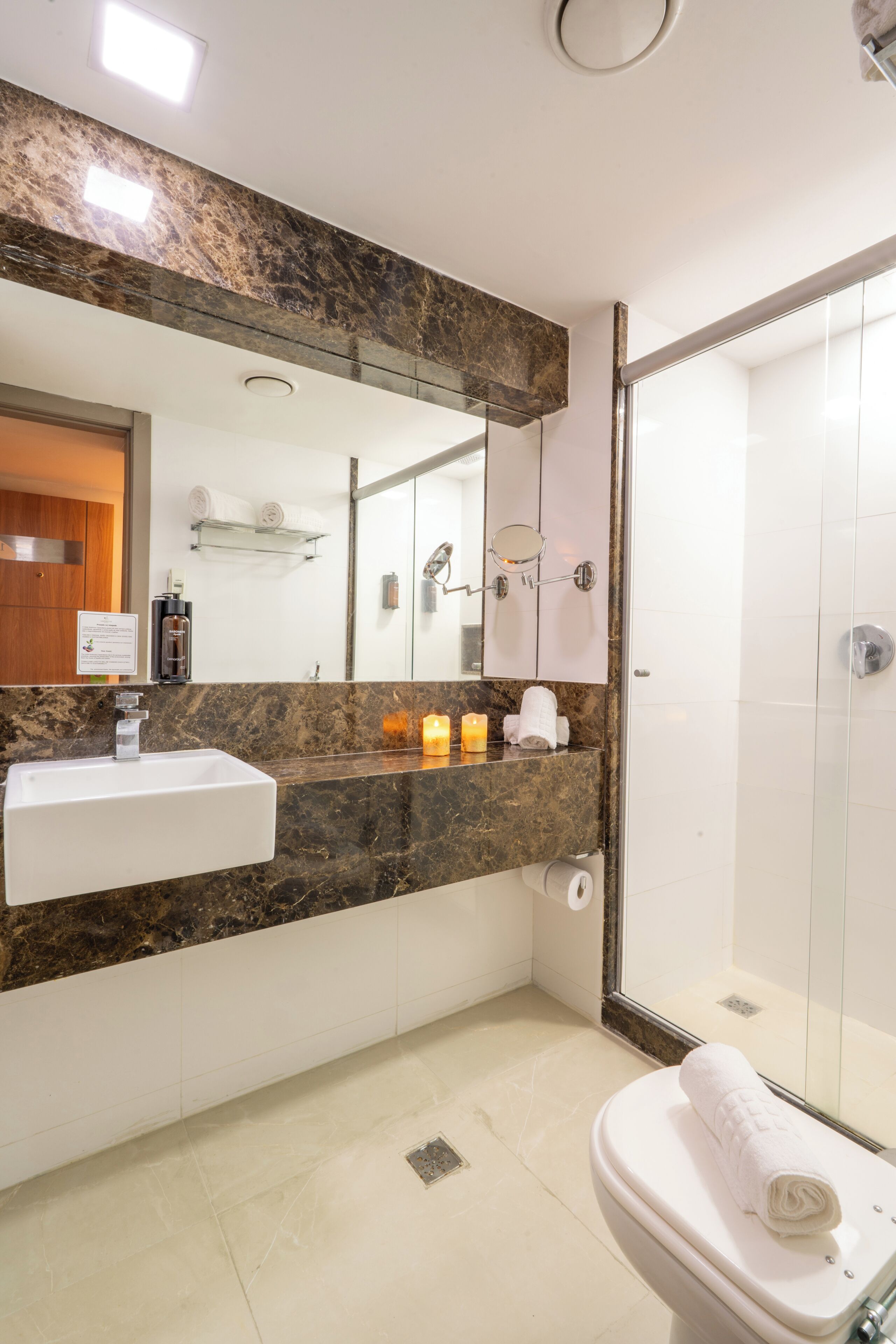 A modern bathroom with a marble countertop, a white sink, a mirror, a glass shower enclosure, and a toilet with a towel.