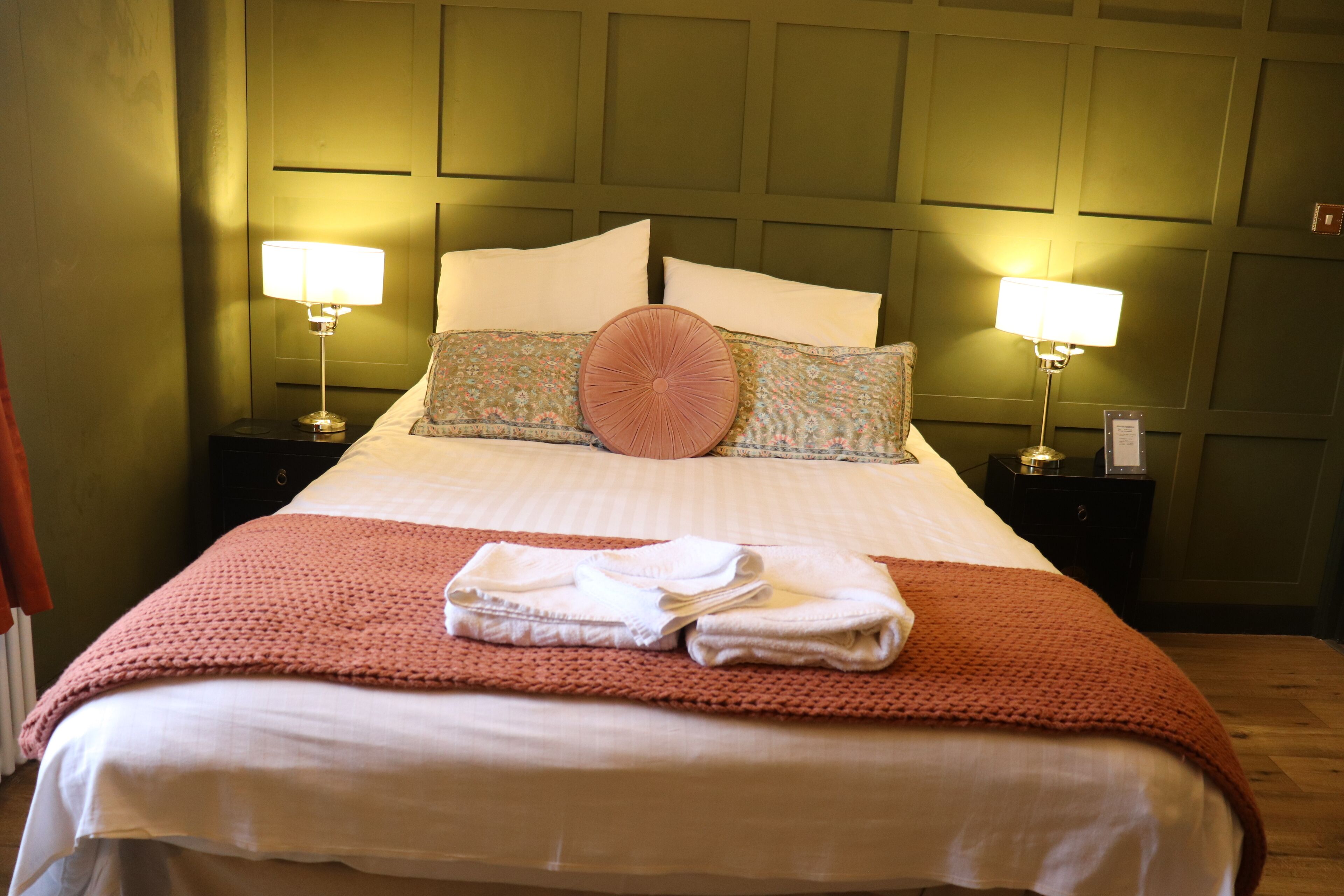 A neatly made bed with a pink knitted blanket, two bedside lamps, and a headboard with a panel design.