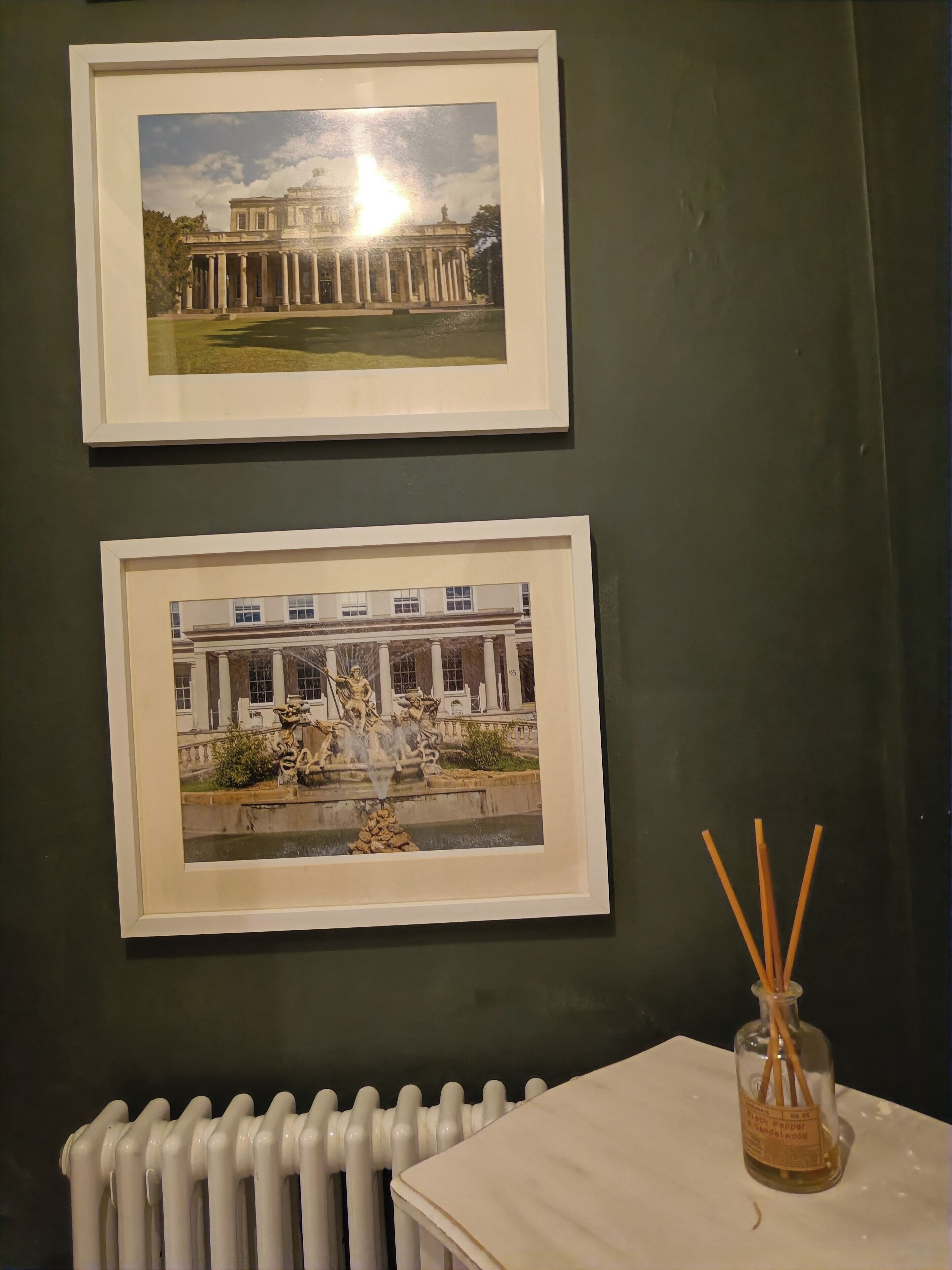 Two framed pictures of classical architecture on a dark wall, a white radiator below, and a glass bottle with reed sticks on a table.