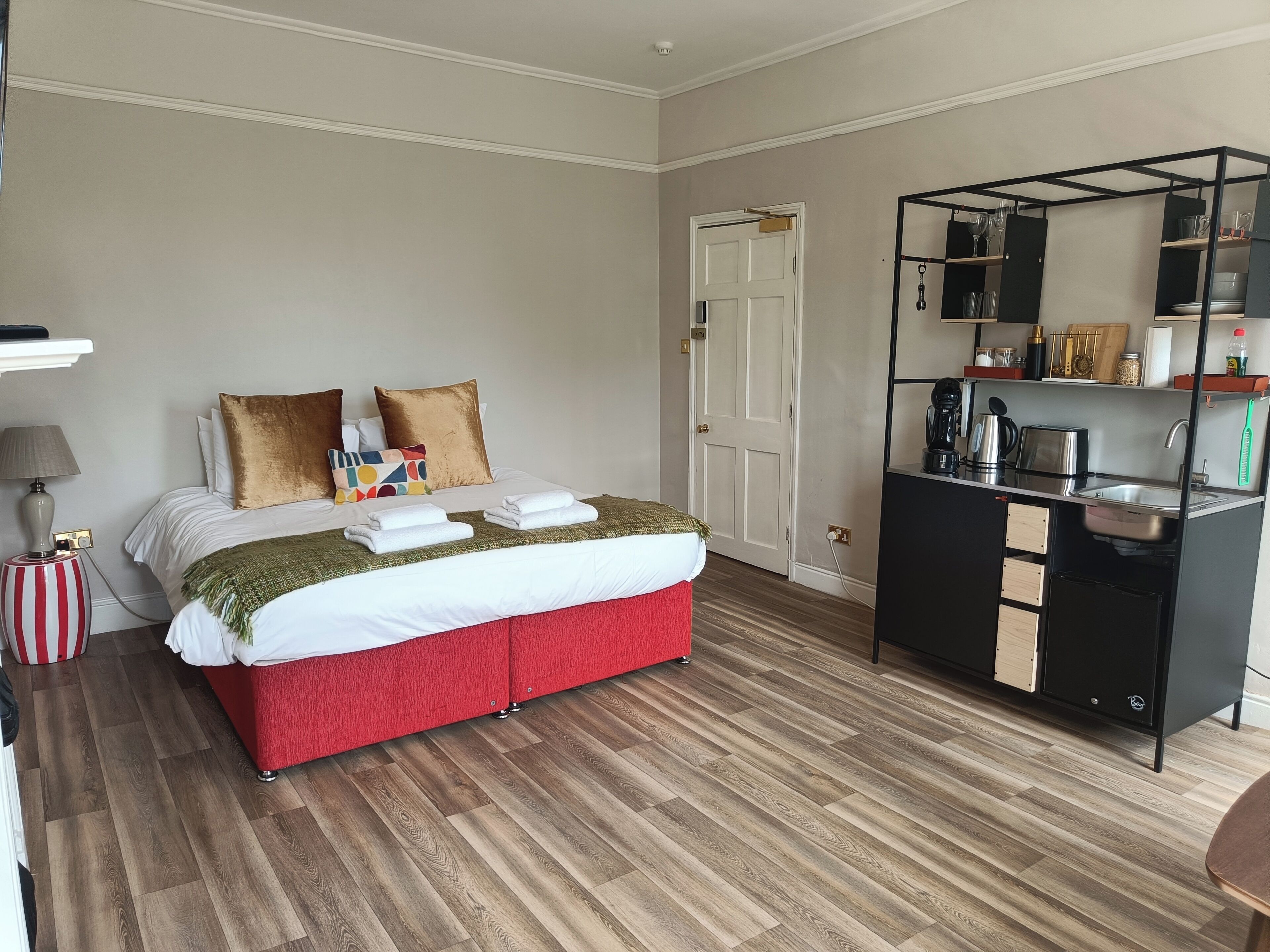 A hotel room with a bed, a bedside table with a lamp, a red ottoman, a kitchenette with a sink and stove, and a door leading to another room.