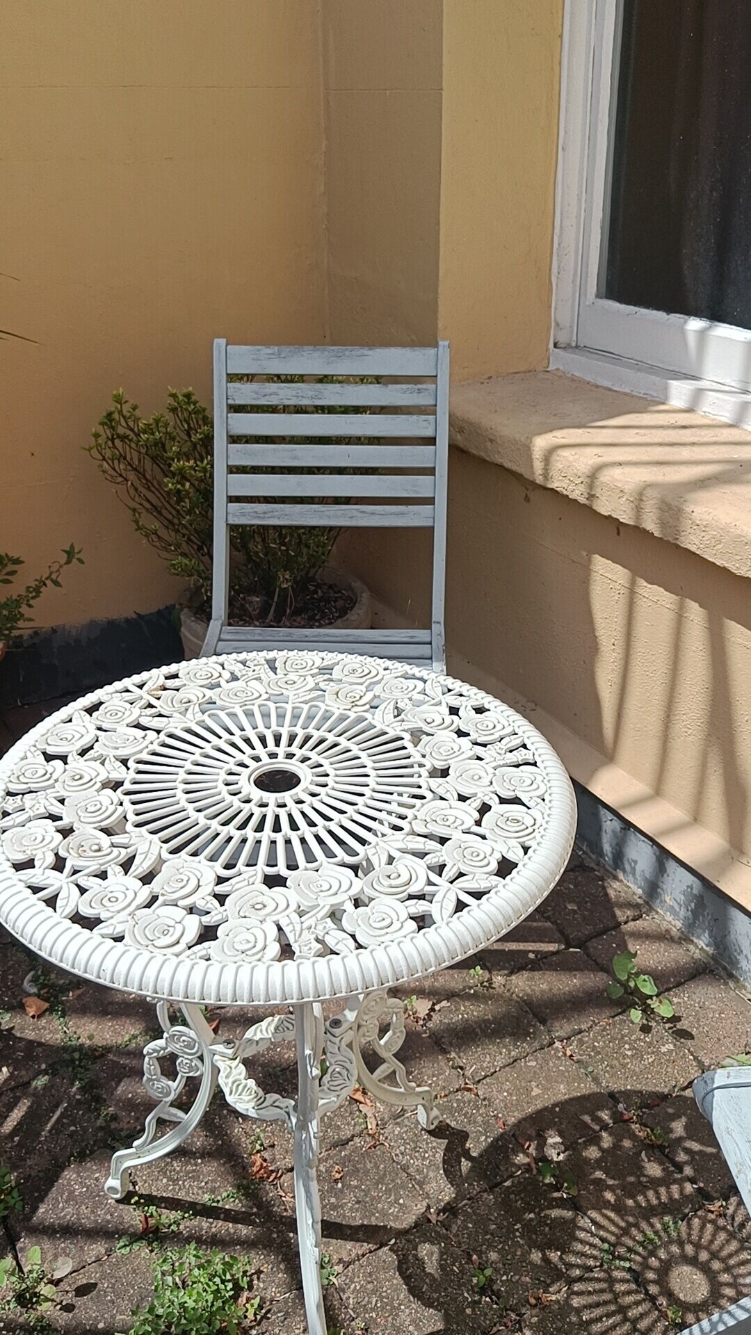 Un tavolo in metallo bianco con elaborate decorazioni floreali, una sedia abbinata e una pianta in vaso in un angolo.