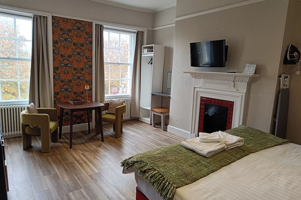 A hotel room with a wooden floor, a bed with a green throw, a fireplace, a TV mounted above it, a dining table with chairs, and a window with curtains.