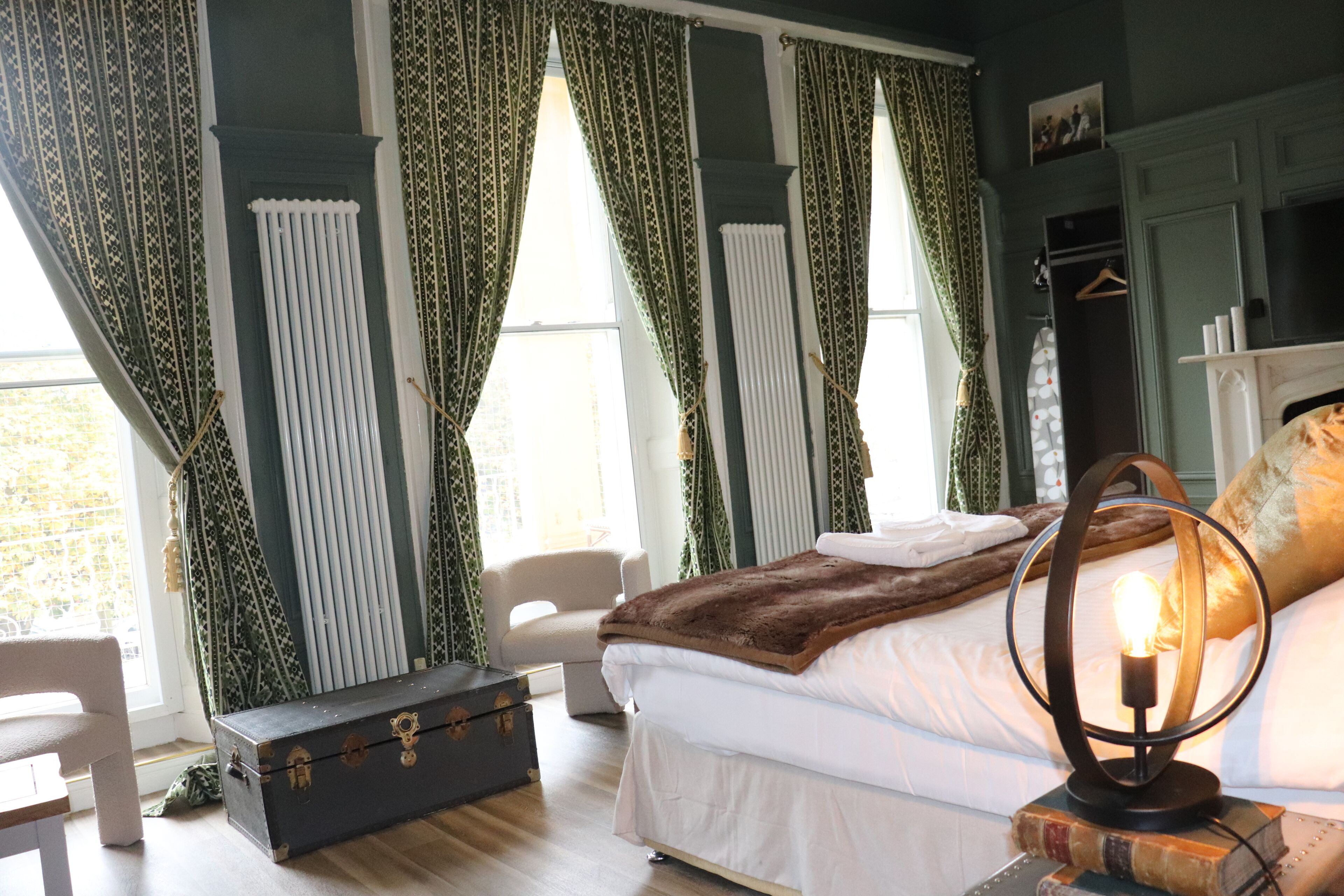 Un dormitorio con cama, maleta, lámpara y ventana con cortinas.