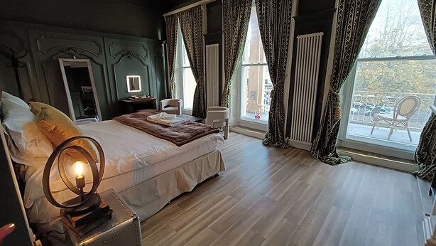 A bedroom with a large bed, a bedside table with a lamp, a chair, and a view of the outdoors through a window. - Rooms on The Prom (Cheltenham)