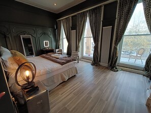 A bedroom with a large bed, a bedside table with a lamp, a chair, and a view of the outdoors through a window. - Rooms on The Prom (Cheltenham)