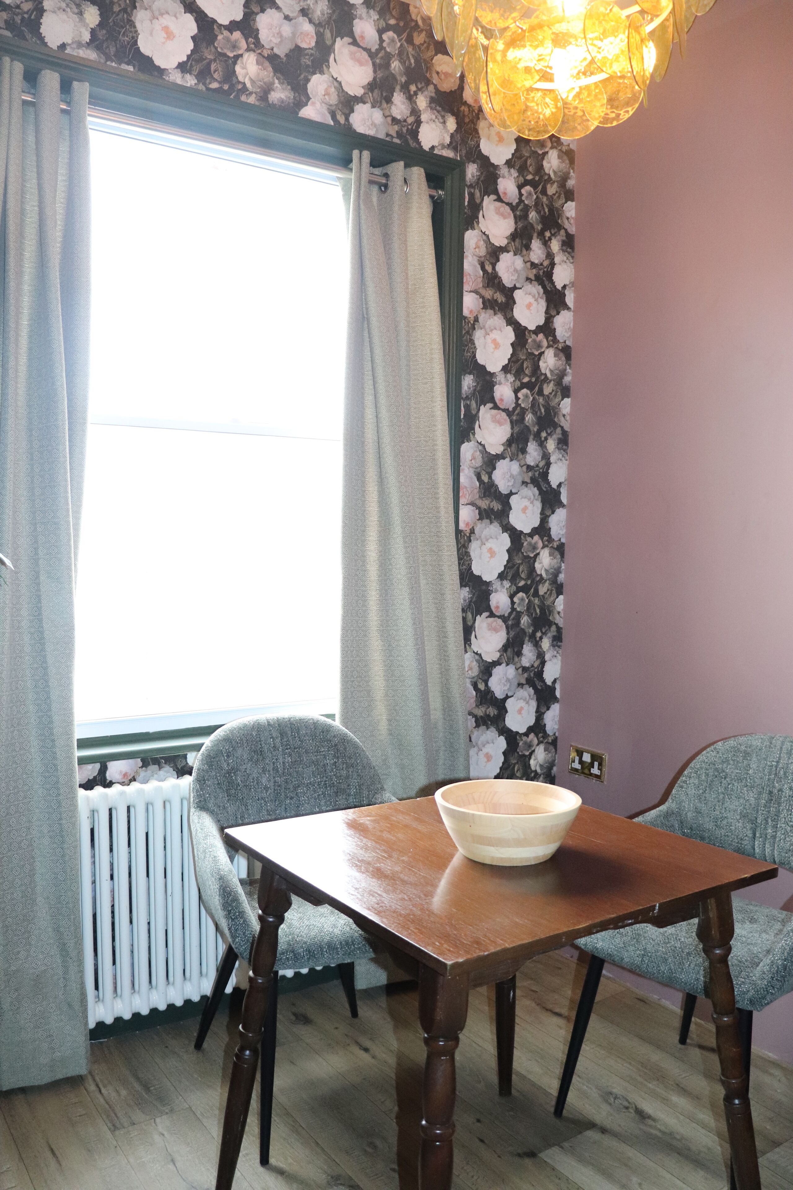 A dining area with a wooden table and two chairs, a window with curtains, a radiator, and a floral wallpaper.